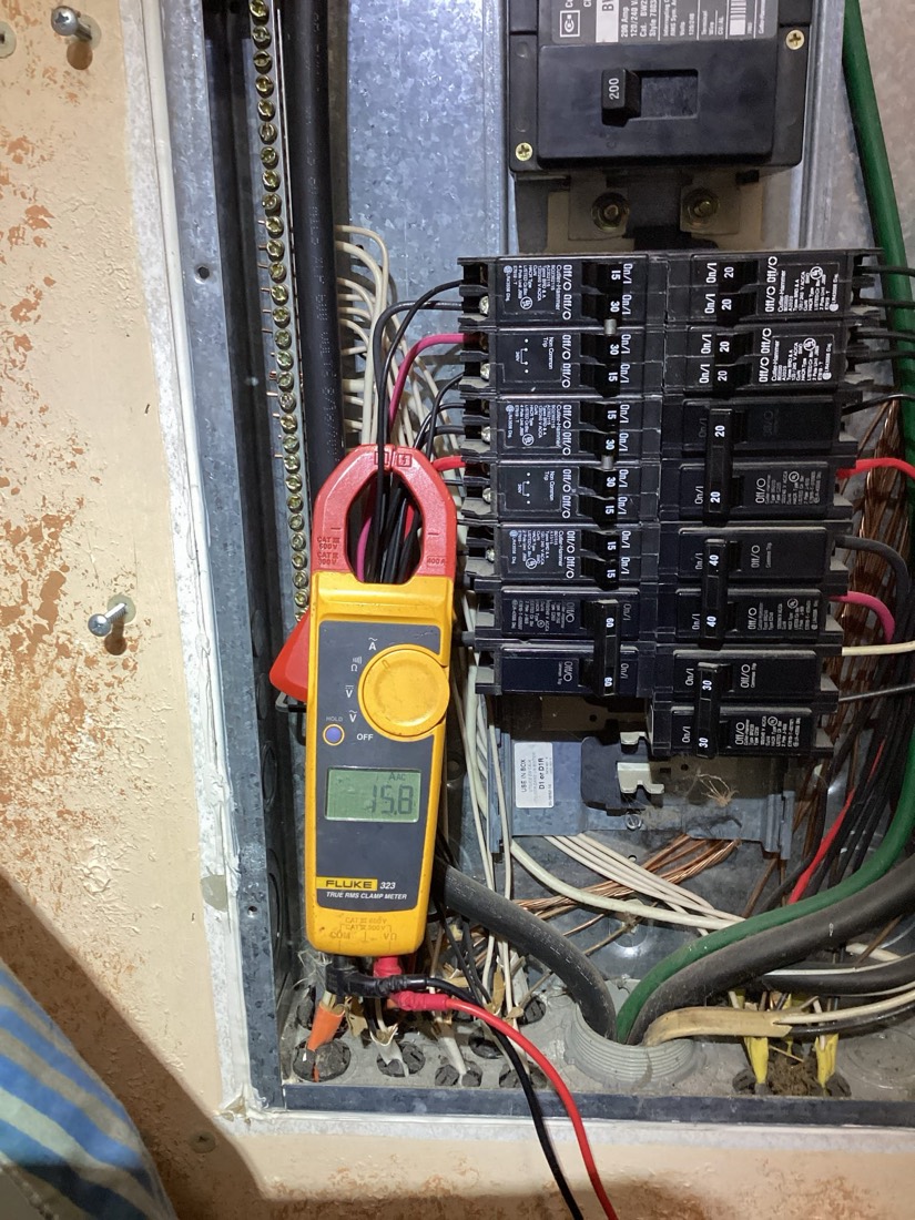 Clamp meter on breaker panel with labeled circuit breakers and wiring.