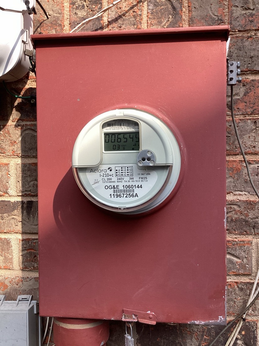 Electric meter on red exterior panel mounted to brick wall.